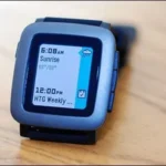 Một chiếc đồng hồ Pebble màu đen với màn hình đơn sắc hiển thị giờ trên nền gỗ.