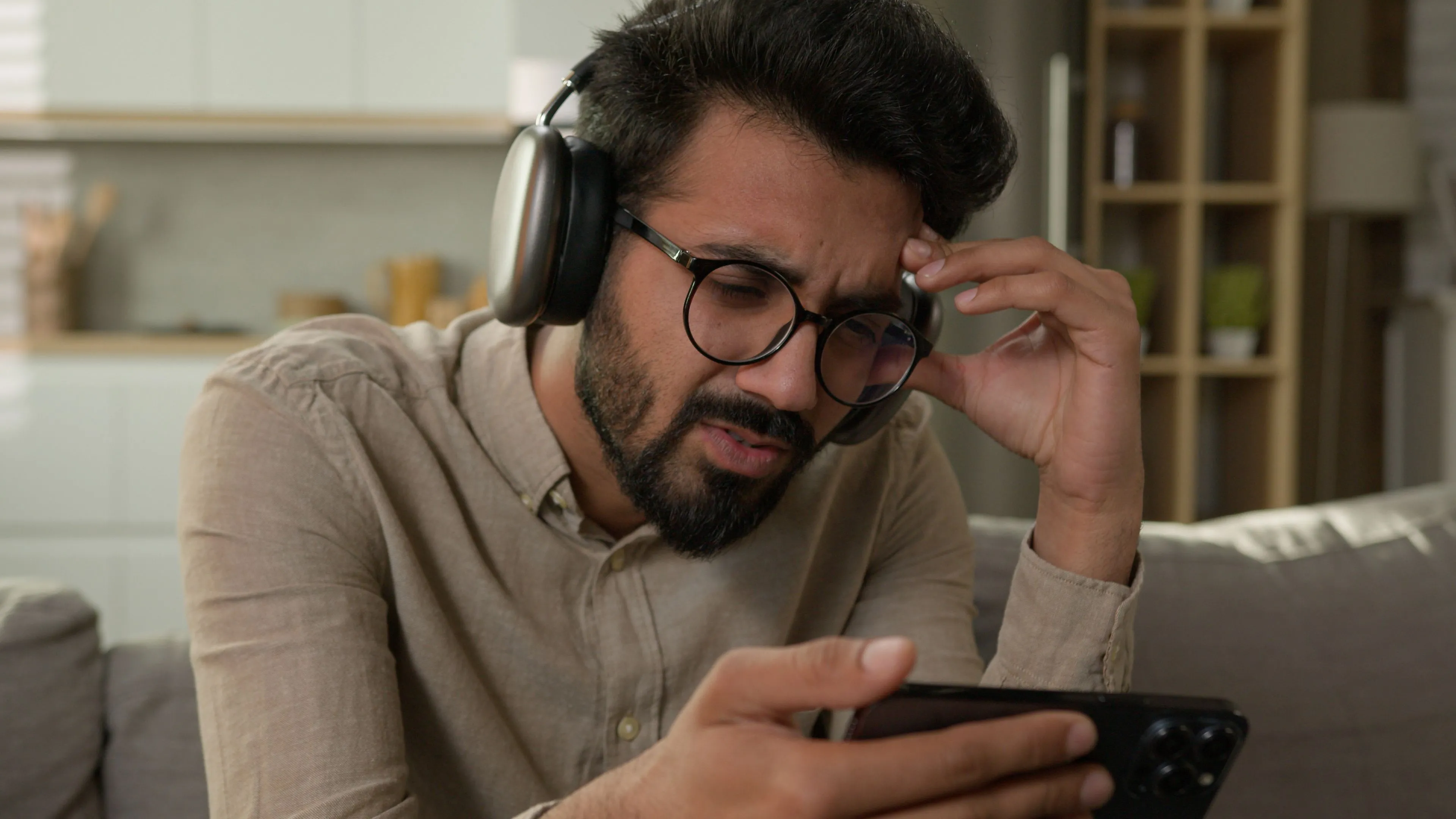 Người đàn ông cau mày nhìn vào iPhone với tai nghe AirPods Max, thể hiện sự mệt mỏi khi chơi game gacha quá mức