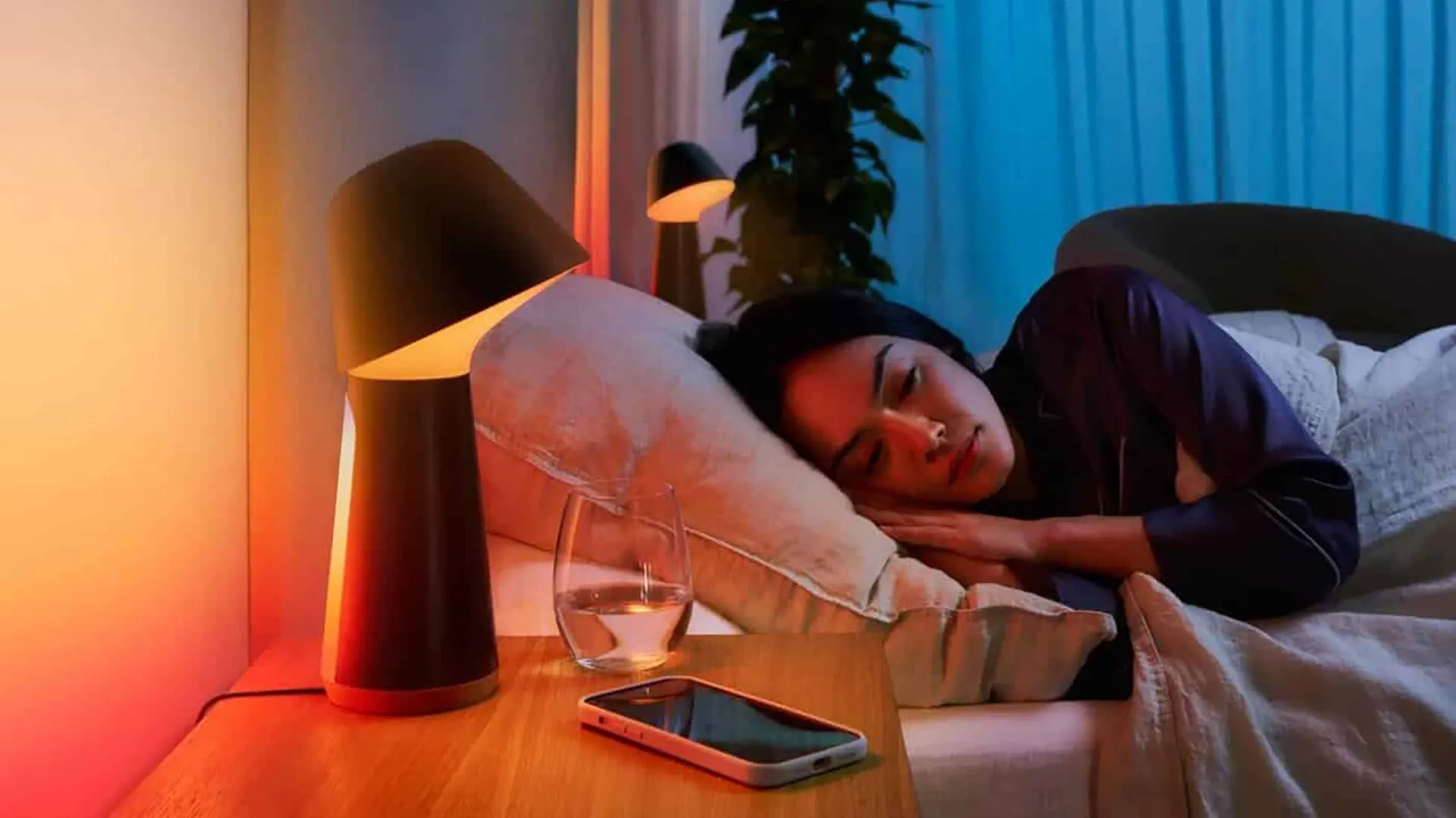 Người phụ nữ đang ngủ trên giường, bên cạnh là đèn ngủ thông minh và điện thoại di động, minh họa việc sử dụng điện thoại để hỗ trợ giấc ngủ.
