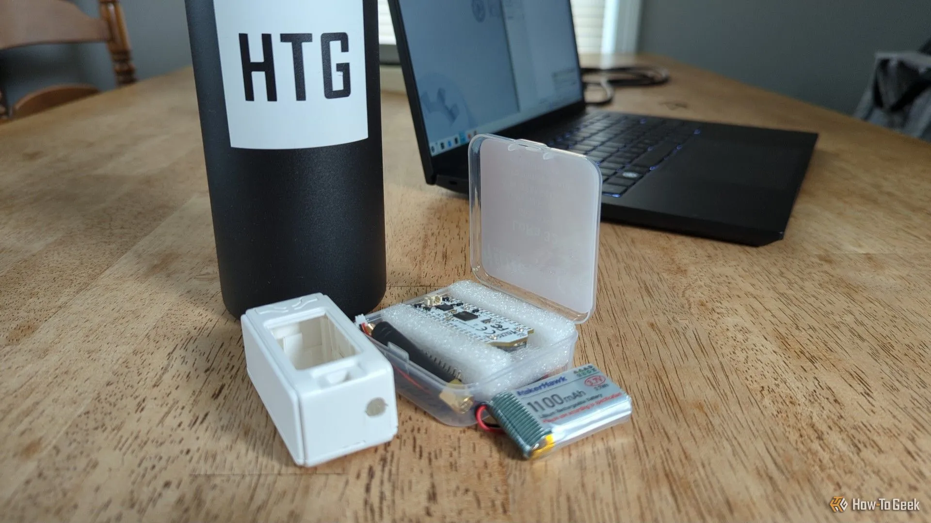 Bộ kit radio LoRa MakerHawk với bảng mạch, vỏ và pin có thể nhìn thấy phía trước laptop và chai nước logo How-To Geek