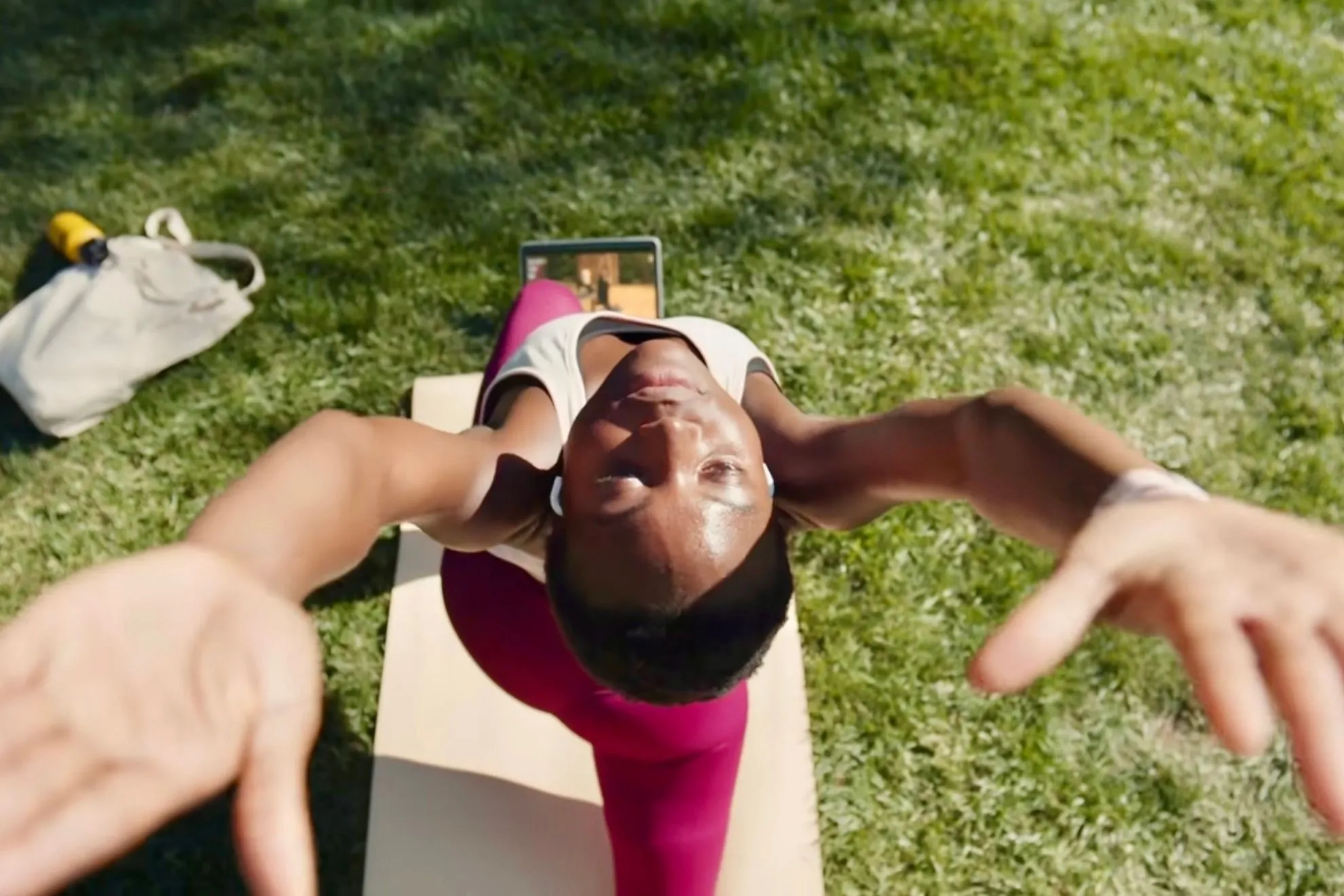 Một người đang sử dụng Apple Fitness+ để tập yoga tại công viên trên bãi cỏ, với thảm yoga và iPad.