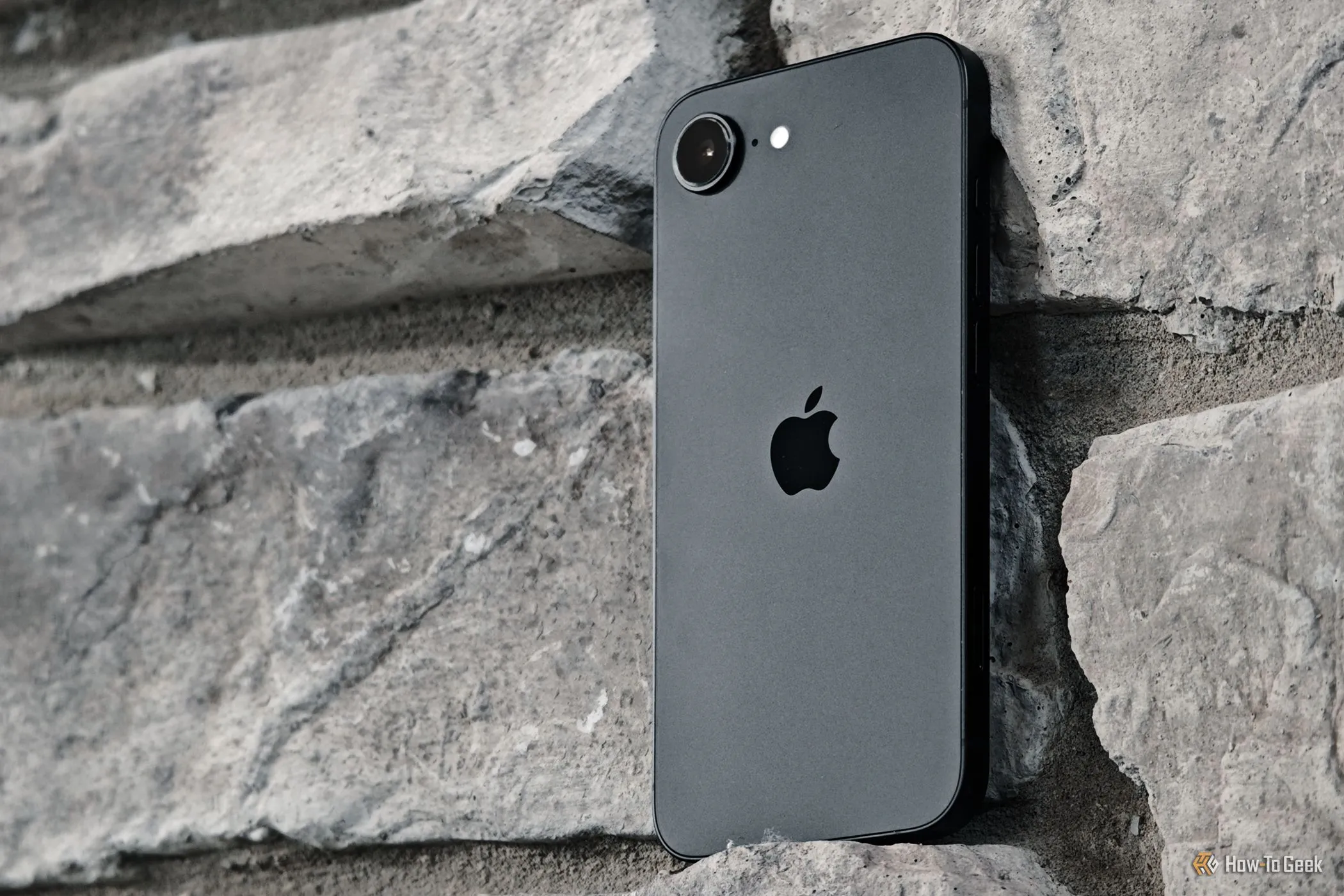 iPhone 16e màu đen nổi bật trên nền tường gạch, phô diễn vẻ đẹp tối giản và khả năng cầm nắm chắc chắn của chiếc điện thoại Apple